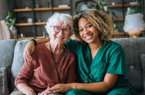 Smiling nurse in green scrubs sitting with cheerful elderly woman in cozy home setting