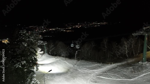 Drone footage of a modern cable car transporting tourists to the mountain peak over snowy ski trails at night.