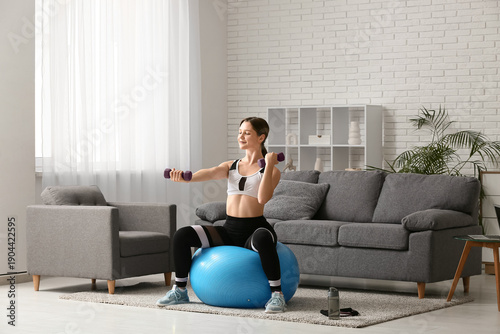 Beautiful young woman training with dumbbells on fitball in living room