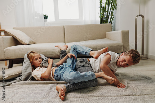 Фотография Two playful children in casual clothes wrestle on the carpet in a bright living room with modern sofa and plants