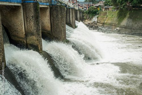 Strong river currents rush over a low katulampa concrete dam, forming white water and foam, showing freshwater flow, simple hydraulic engineering, and natural movement, landscape view