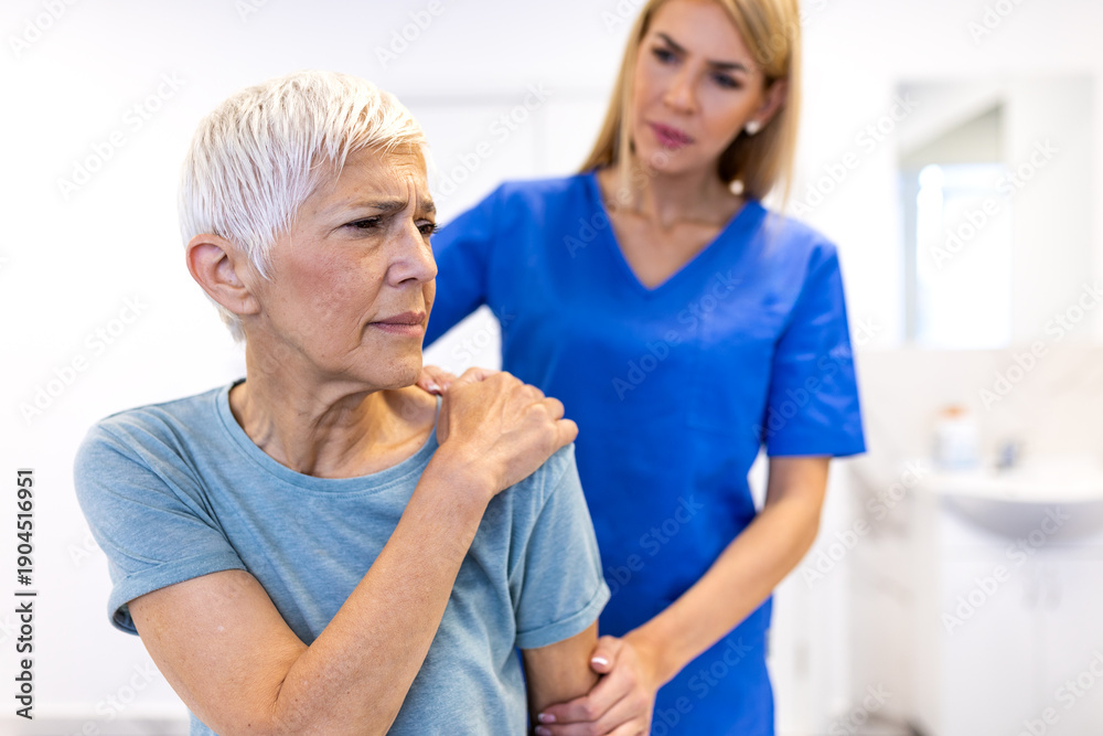 Obraz premium Pain in the shoulder. Upper arm pain, people with body-muscles problem, healthcare and medicine concept. Senior woman sitting in clinic and holding painful shoulder while nurse or doctor helps her.