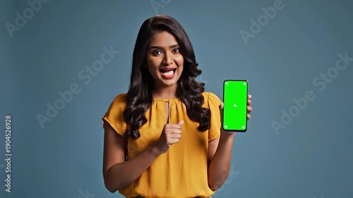 Smiling young Indian woman in a bright yellow top pointing at a smartphone with a vibrant green screen for easy copy space and advertising mockups in studio settings.