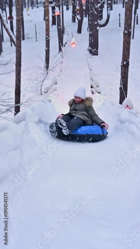 A young girl in warm winter attire slides down snowy hills on a blue snow tube. Laughter fills the air as she enjoys the thrill of speed among tall trees in a serene winter landscape.