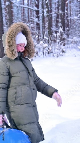 A young girl walks through a winter wonderland, pulling her bright blue snow tube along the snowy path.
