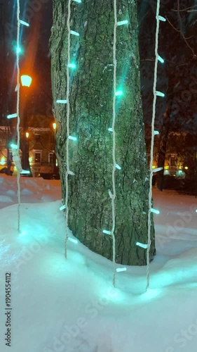 The vertical video shows a city street at night, where garlands on trees, lampposts, and buildings shimmer with multicolored lights through the fresh snow. 