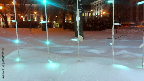 The vertical video shows a city street at night, where garlands on trees, lampposts, and buildings shimmer with multicolored lights through the fresh snow. 