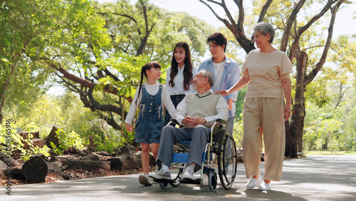 An Asian family enjoys a walk in a lush green park, supporting a senior man in a wheelchair. Multigenerational family authentic love, care, accessibility, and happiness outdoors