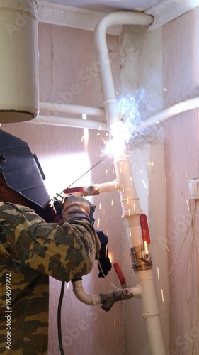 A welder, seen from behind, wearing a uniform and hard hat, welds a section of a house's heating system, made of white-painted iron pipes, to the ceiling.