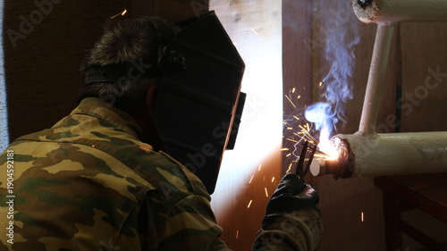A welder, seen from behind, in uniform and a protective helmet, welds a section of a home heating system made of white-painted iron pipes. A man wearing a special mask welds a heating radiator.