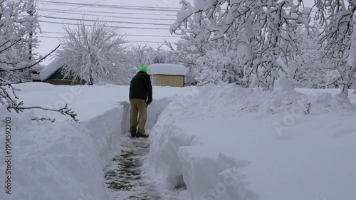 A man in warm clothing, seen from behind, clears a path through high snowdrifts on a residential property leading to a roadway in a rural area. Clearing a path leading from a house to a road