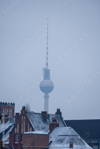 Berlin TV Tower rises above snowy rooftops in winter haze, its sphere and antenna fading into the cold gray sky over the quiet cityscape
