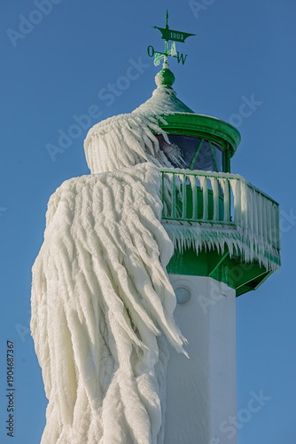 Detailaufnahme: Vereister Leuchtturm Sassnitz mit Eisvorhang und Eiszapfen am grünen Laternenhaus vor blauem Himmel, Rügen, Ostsee