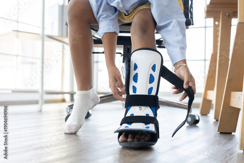 Foto Child sits on chair while wearing an orthopedic ankle brace, adjusting medical foot support for injury recovery, mobility assistance, pediatric healthcare, rehabilitation treatment at home