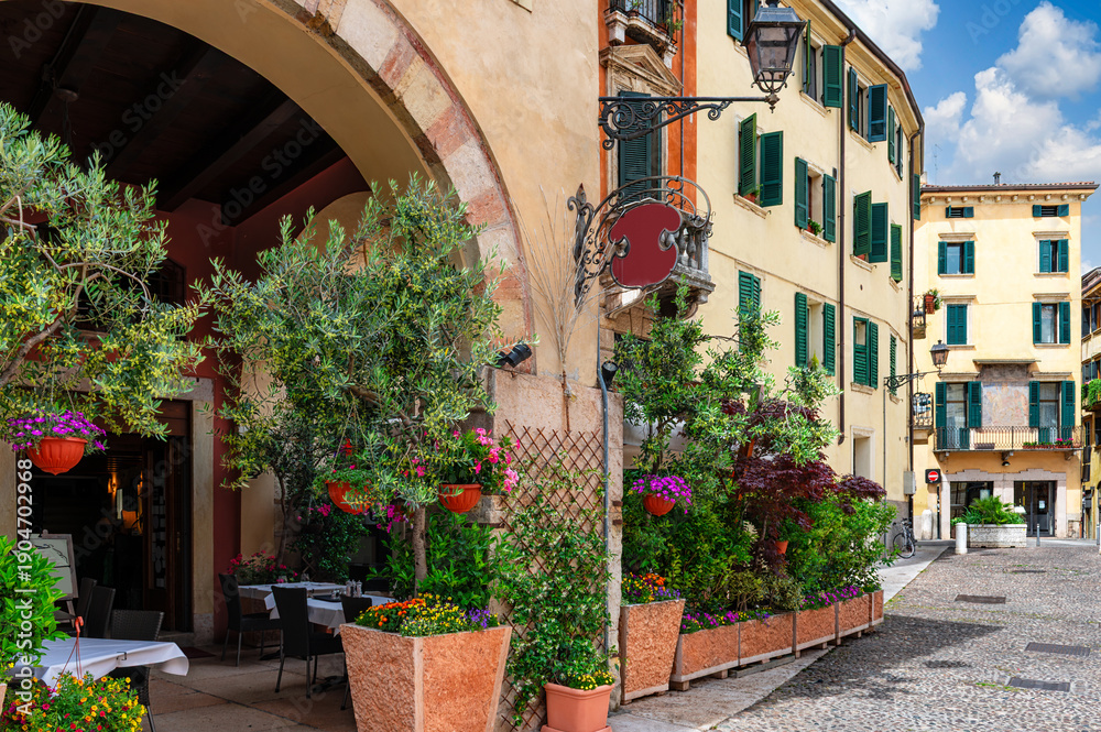 Obraz premium Old street with flowers and tables of restaurant in Verona, Italy. Architecture and landmark of Verona. Cozy cityscape of Verona.
