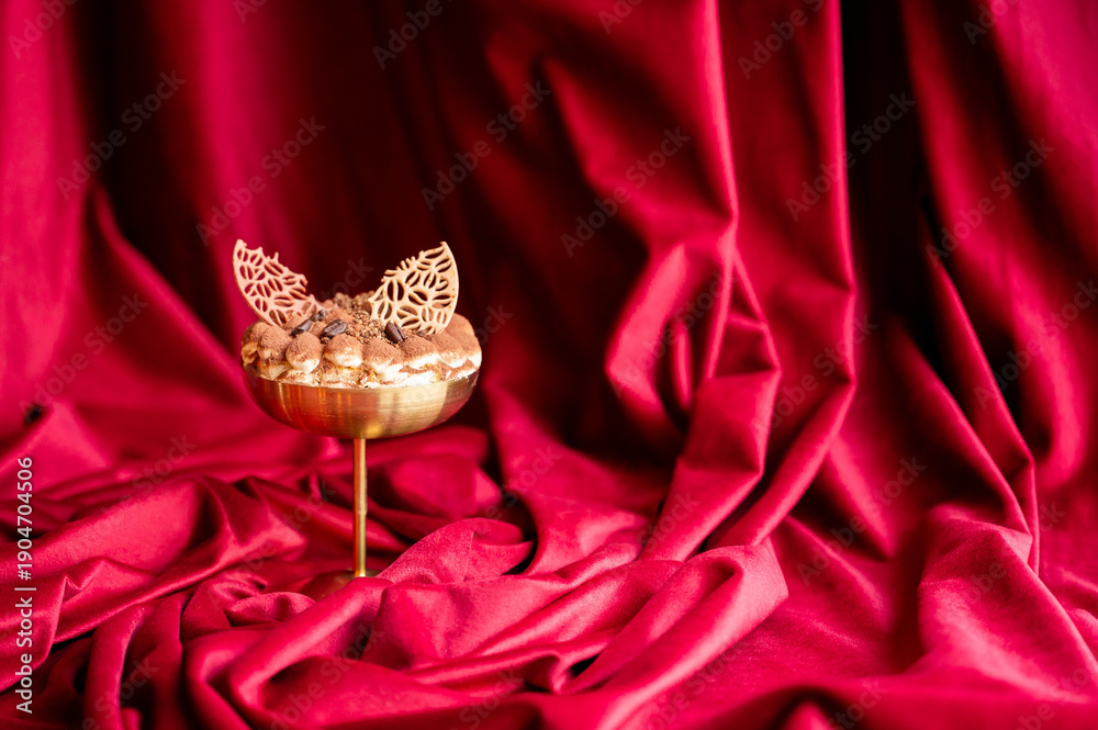 Obraz premium Elegant gourmet tiramisu dessert served in a gold coupe glass, topped with chocolate coffee bean garnishes and cocoa powder, set against a luxurious, draped red velvet fabric background