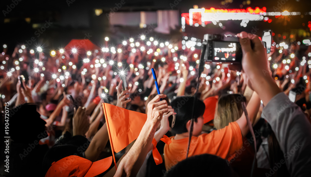 Fototapeta premium Close up of a hand holding a pen ready for voting at a People's Party rally in Thailand. Supporters with orange flags and phone lights in background for February 8, 2026 election.