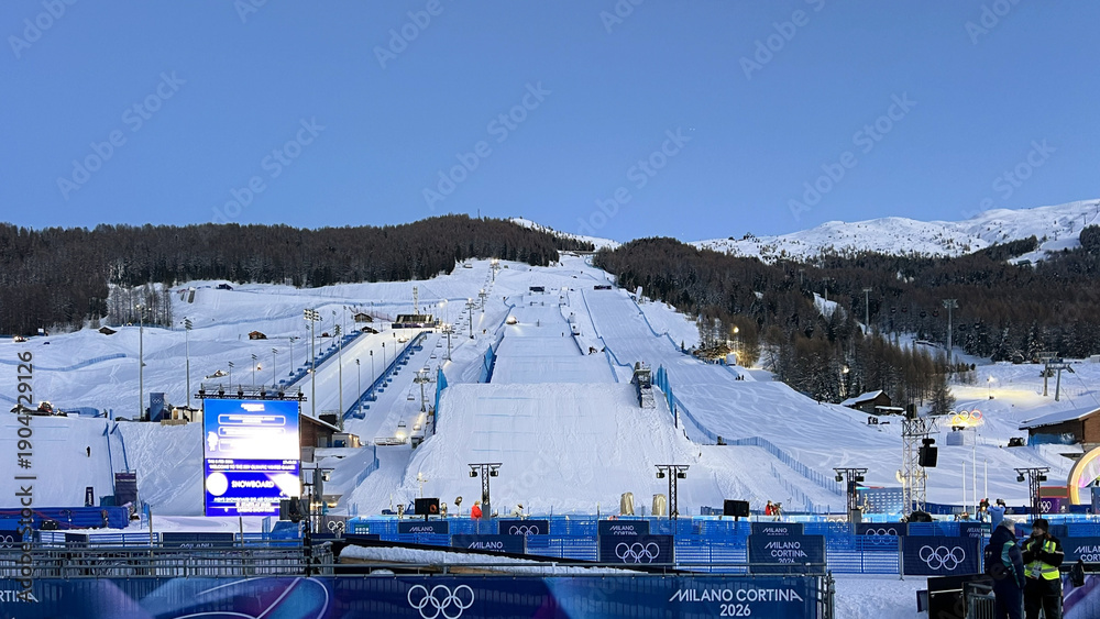 Naklejka premium Livigno, Italy - February 6, 2026: Freestyle skiing venue with massive snow jump and competition course prepared for Winter Olympic Games in Livigno under clear blue sky