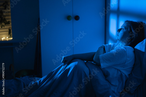 Принти на полотні Exhausted middle-aged woman sitting in bed and looking through the window at nig