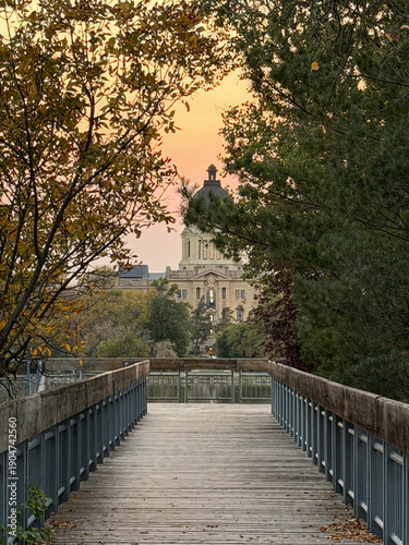 A wooden path leads through autumn trees to the Saskatchewan Legislative Building in Regina, glowing under a colorful sunset sky.