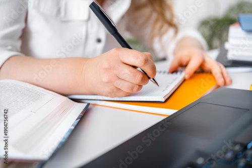 Tablou pe pânză Close up of woman hand writing notes in notebook near open book and laptop, adul