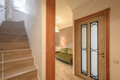 Wooden staircase with light steps, warm hallway with wooden door featuring frosted glass panels, green sofa in background, soft lighting, neutral tones.