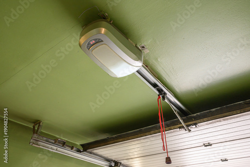 A ceiling-mounted garage door opener with a metallic casing, control panel, and red emergency release cord. Installed above a sectional garage door, fixed to a green ceiling
