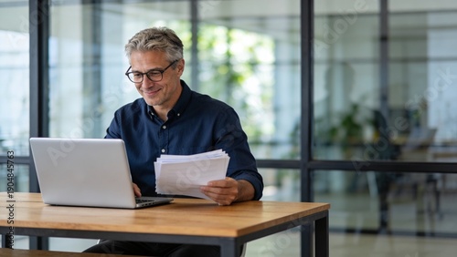 A man is sitting at a desk with a laptop and a stack of papers