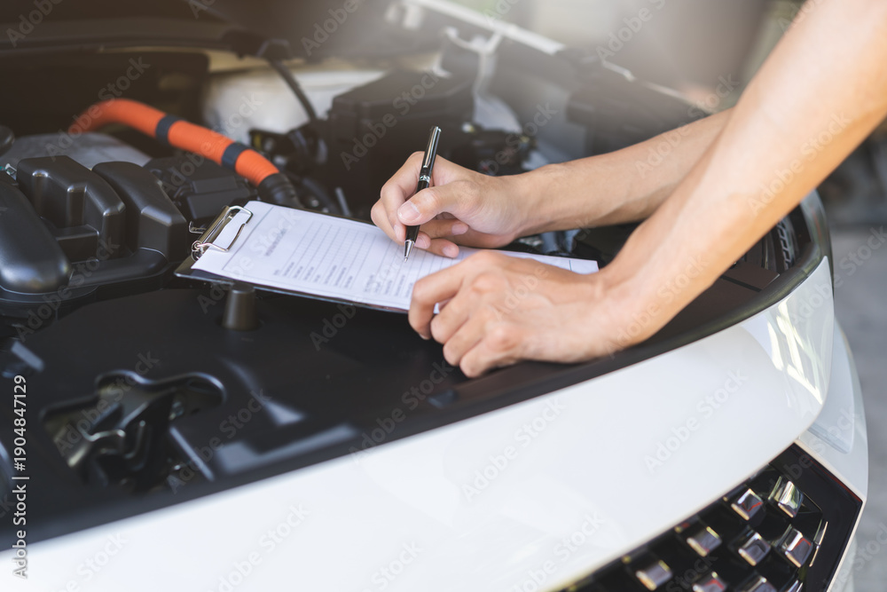 Fototapeta premium Auto check, car service shop concept. Automobile repairman writing job checklist on clipboard, mechanic checking engine to estimate repair machine, inspecting maintenance by engineer at vehicle garage