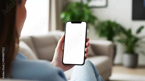Hand holding mobile smartphone with blank white screen mockup while woman sitting in modern living room interior background for app design website display copy space branding marketing
