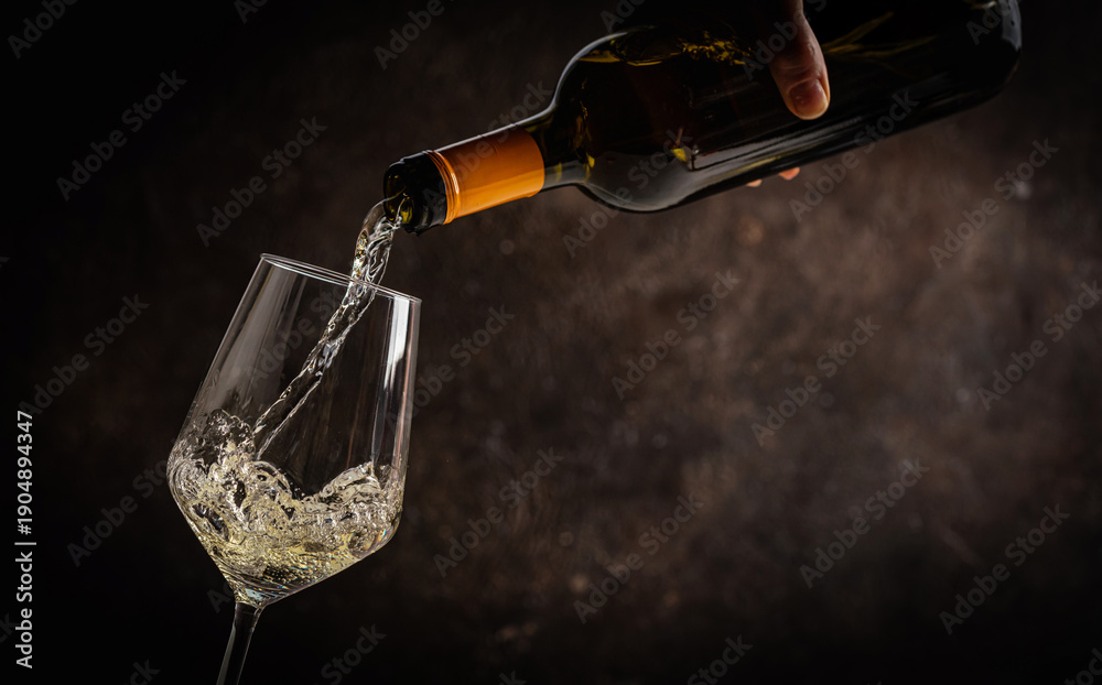 Fototapeta premium Pouring white wine into the glass on rustic dark background. Pour alcohol, winery concept.