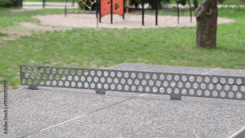 Close-up of a ping pong ball in mid-air over a concrete table tennis table in a park. Players passing the ball back and forth, surrounded by green trees and grass. Captures dynamic motion and outdoor 