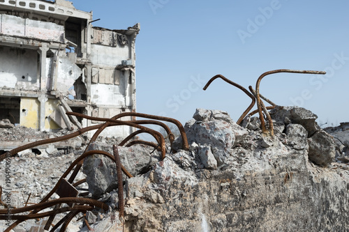 Destroyed concrete structure with exposed rusty metal rebar under blue sky, remains of demolished building and rubble. Background.