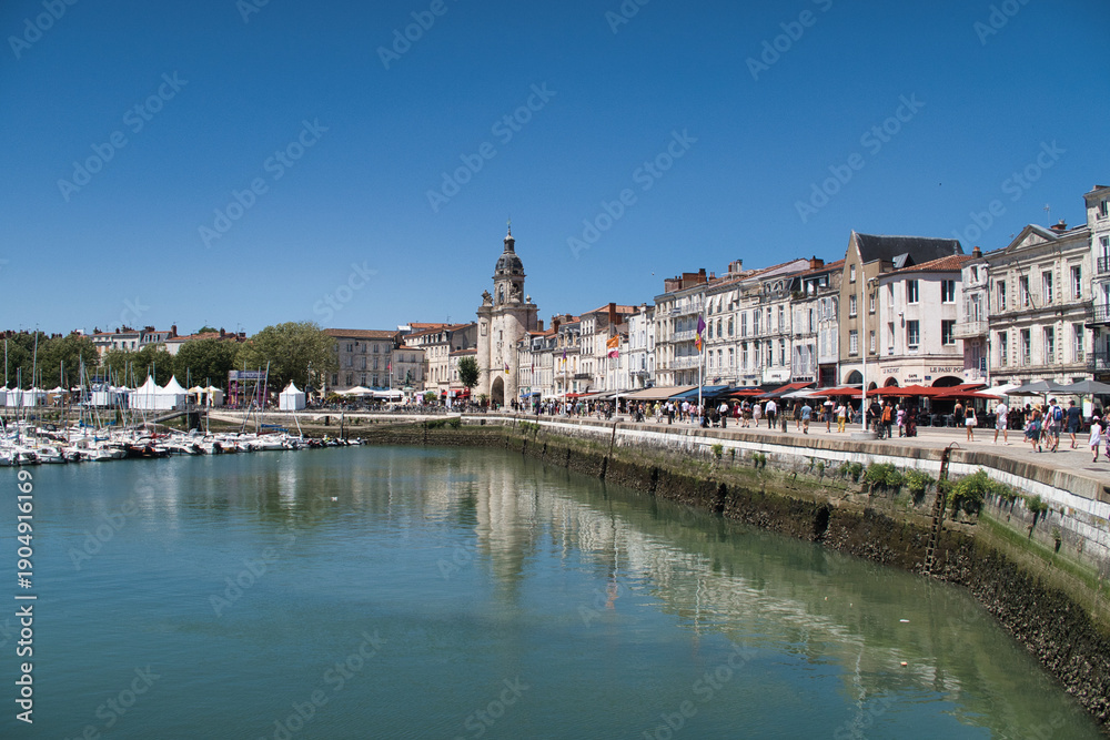 Obraz premium Historic European Port City View - Wide view of a sunny French port with historic waterfront buildings, a prominent clock tower, and boats reflected in the calm water.