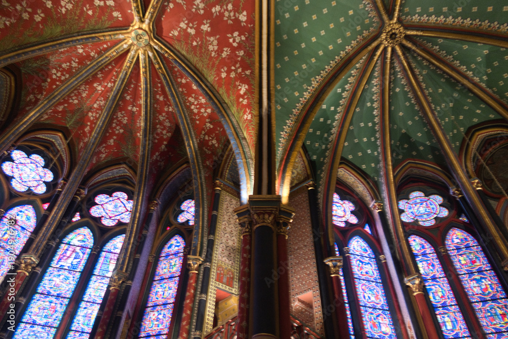 Obraz premium Gothic Vaults and Murals of Sainte-Chapelle - Low-angle view of richly painted Gothic rib vaulting in red, gold, and teal, alongside a large crucifixion mural and stained glass windows.