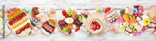 Spring or Easter grazing board theme table scene against a white wood banner background. Overhead view. Selection of sweets and cheese, meat and fruit appetizers.