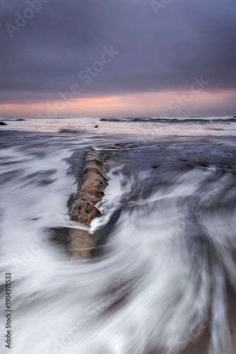 Fotografi Long exposure of sea waves flowing around a rock on Azkorri beach under a dark c