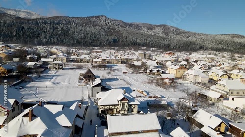 Houses in a village near snow-covered mountains with pine trees in winter. Aerial panoramic drone view from above, top view from drone.  Ukraine, Carpathians, Skole city
