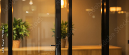 Modern office interior viewed through glass door with black metal frame, blurred background with plants and warm lighting, corporate workspace concept for architectural design and business background