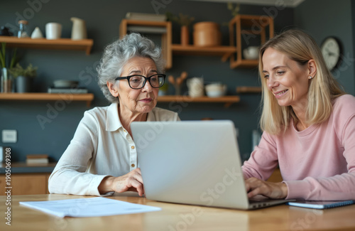 Elderly woman learns using laptop with younger woman assisting. Two females interact at table, sharing digital device for online activity. Mature lady types on computer keyboard with help from adult