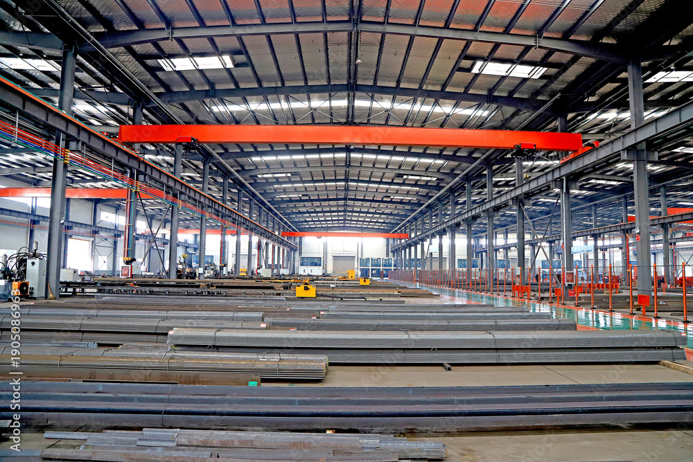 Obraz premium Spacious Steel Processing Factory Workshop with Overhead Cranes and Stacked Steel Profiles