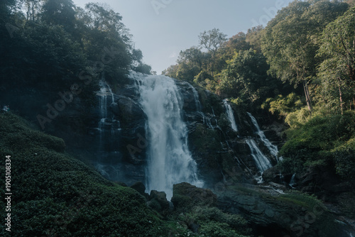 A majestic waterfall cascades down a mossy cliffside, surrounded by lush green vegetation and bathed in soft morning light.
