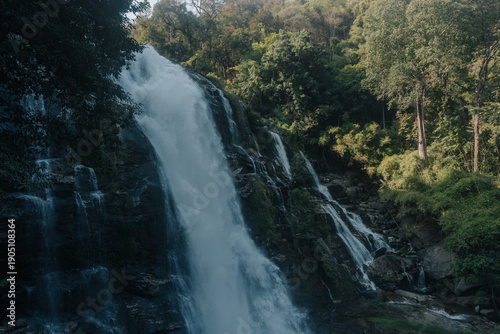 A powerful waterfall cascades down a mossy cliffside, surrounded by lush green forest and bathed in soft, golden light.