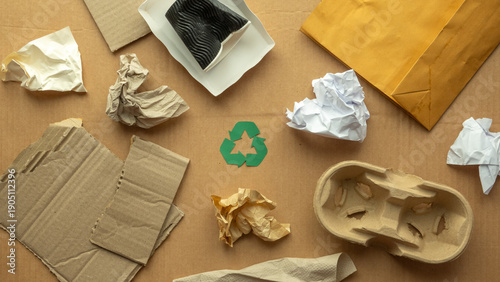 Flat lay of recyclable paper packaging waste with cardboard, envelopes, crumpled paper and green recycle symbol on brown background, eco friendly lifestyle, recycling and zero waste concept.