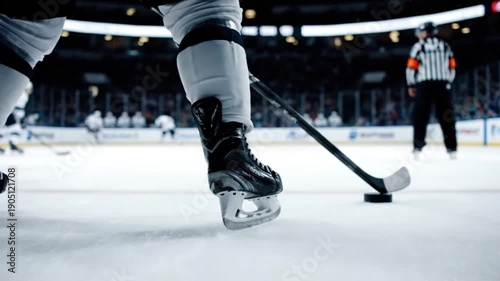 Close-up of an ice hockey player's skate and stick on the ice during a game, with a puck nearby and a referee in the background.
