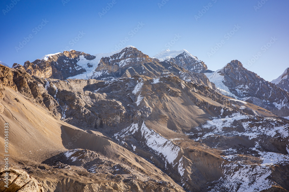 Obraz premium Breathtaking view of a rugged Himalayan mountain range featuring snow-dusted slopes, sharp rocky peaks, and layered alpine terrain beneath a clear blue sky. The vast, untamed landscape