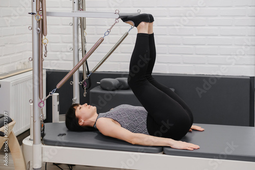 Woman training on pilates tower apparatus with resistance cables, performing advanced core and leg strengthening exercise in contemporary studio