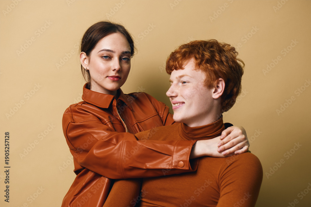 Obraz premium Young couple with brown hair and casual outfits embracing each other on beige background. Woman with leather jacket holding smiling redhead man in close pose.