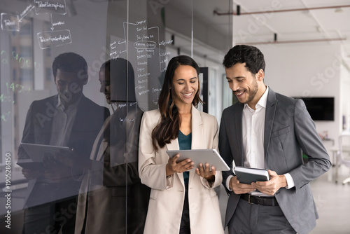 Two smiling teammates, male, female start-up members using tablet stands next to glass black board with written notes, ideas or strategy plan. Teamwork, modern technology, business application usage