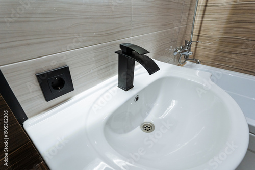 Matte Black Faucet Over Ceramic Sink, Compact Vanity With Dark Outlet And Tiled Wall, Soft Reflections And Clean Lines Convey Contemporary Bathroom Styling And Minimal Design